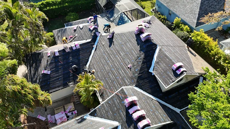 Aerial view of a full shingle roof replacement in progress, with synthetic underlayment installed, shingle bundles staged across the deck and the crew working beside a screened pool